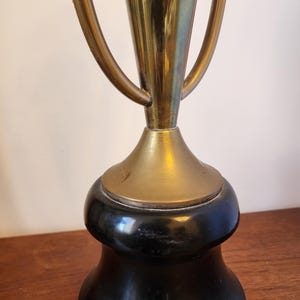Pode incluir: Um trof&eacute;u com uma ta&ccedil;a de metal em tom dourado com duas al&ccedil;as curvas, montado sobre uma base preta lisa e alargada. A sec&ccedil;&atilde;o dourada tem um acabamento polido, enquanto a base &eacute; de cor preta s&oacute;lida com um aspeto arredondado e pesado.
