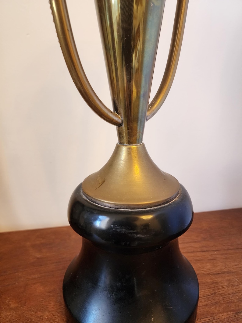 Pode incluir: Um trof&eacute;u com uma ta&ccedil;a de metal em tom dourado com duas al&ccedil;as curvas, montado sobre uma base preta lisa e alargada. A sec&ccedil;&atilde;o dourada tem um acabamento polido, enquanto a base &eacute; de cor preta s&oacute;lida com um aspeto arredondado e pesado.