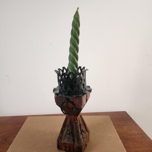 May include: A dark green spiral taper candle sits inside a black metal crown-shaped holder, which is mounted atop a square, weathered brown wooden base featuring a small metal ring and decorative metal studs on its sides.