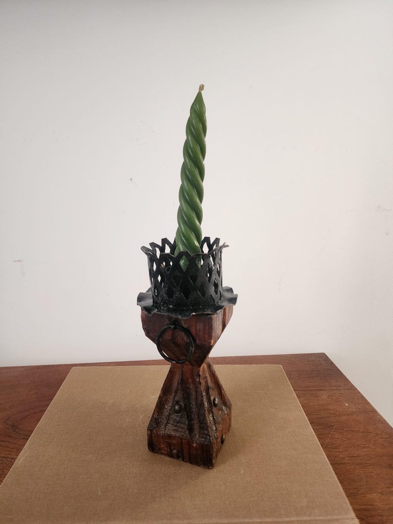May include: A dark green spiral taper candle sits inside a black metal crown-shaped holder, which is mounted atop a square, weathered brown wooden base featuring a small metal ring and decorative metal studs on its sides.