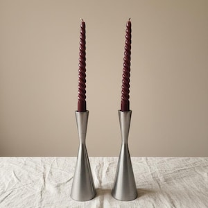 Puede incluir: Dos candelabros de metal cepillado con velas en espiral burdeos. Los candelabros tienen una base acampanada y un cuello delgado. Las velas son de color rojo oscuro y tienen un diseño retorcido. El fondo es de color beige neutro.