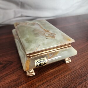 Puede incluir: Una caja rectangular hecha de piedra &oacute;nix veteada en verde claro y blanco. La caja cuenta con una tapa con bisagras, un borde de metal dorado y cuatro peque&ntilde;os pies decorativos dorados. Una etiqueta en el frente dice ONYX OF PAKISTAN MADE IN ITALY.