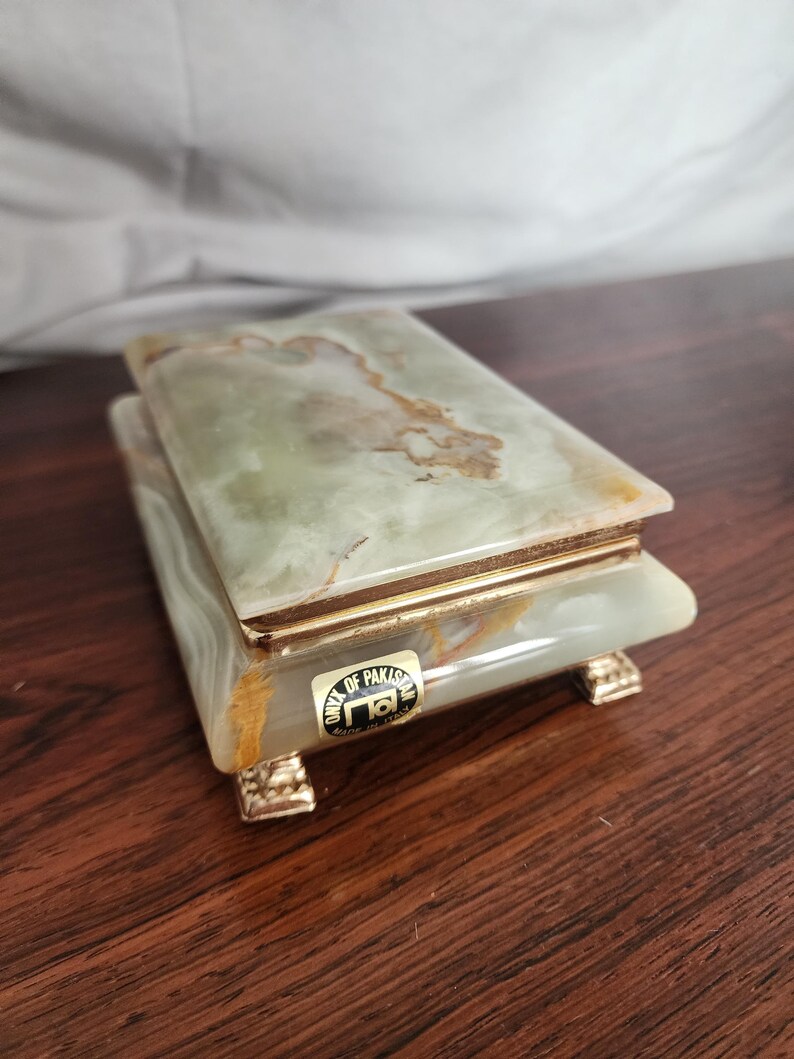 Puede incluir: Una caja rectangular hecha de piedra &oacute;nix veteada en verde claro y blanco. La caja cuenta con una tapa con bisagras, un borde de metal dorado y cuatro peque&ntilde;os pies decorativos dorados. Una etiqueta en el frente dice ONYX OF PAKISTAN MADE IN ITALY.