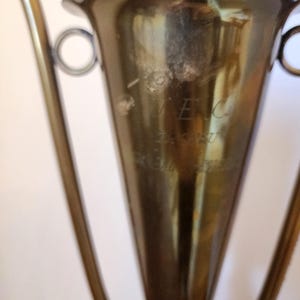 Pode incluir: Uma ta&ccedil;a de trof&eacute;u em metal dourado com corpo c&oacute;nico, rebordo largo e duas pegas laterais curvas. A superf&iacute;cie da ta&ccedil;a apresenta um texto gravado t&eacute;nue que diz VEC, ZAGREB e KEMO-STIL, com sinais vis&iacute;veis de desgaste e oxida&ccedil;&atilde;o no metal.