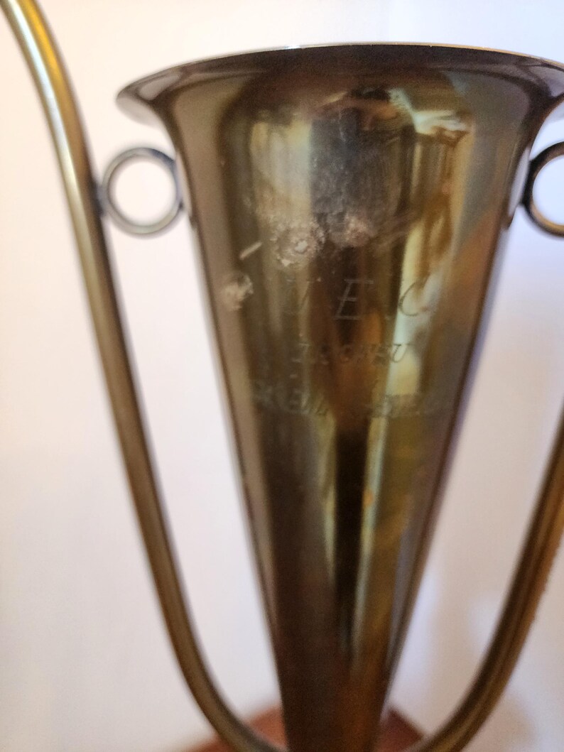 Pode incluir: Uma ta&ccedil;a de trof&eacute;u em metal dourado com corpo c&oacute;nico, rebordo largo e duas pegas laterais curvas. A superf&iacute;cie da ta&ccedil;a apresenta um texto gravado t&eacute;nue que diz VEC, ZAGREB e KEMO-STIL, com sinais vis&iacute;veis de desgaste e oxida&ccedil;&atilde;o no metal.