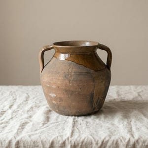 Può includere: Un'antica brocca in ceramica rustica con due manici. La brocca ha un corpo arrotondato e un'ampia apertura. La parte superiore è marrone scuro, che sfuma in un marrone più chiaro nella metà inferiore. Poggia su una superficie strutturata bianco sporco.