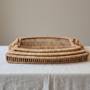 May include: Three woven rattan trays stacked on top of each other. The trays are rectangular with rounded corners and built-in handles. The natural color of the rattan is light brown.