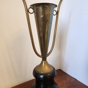 Pode incluir: Uma ta&ccedil;a trof&eacute;u de lat&atilde;o alta com um corpo c&oacute;nico e duas pegas longas curvadas para cima, fixadas a an&eacute;is circulares. O recipiente met&aacute;lico assenta numa base preta lisa e arredondada, criando um contraste entre o metal dourado e o pedestal escuro.