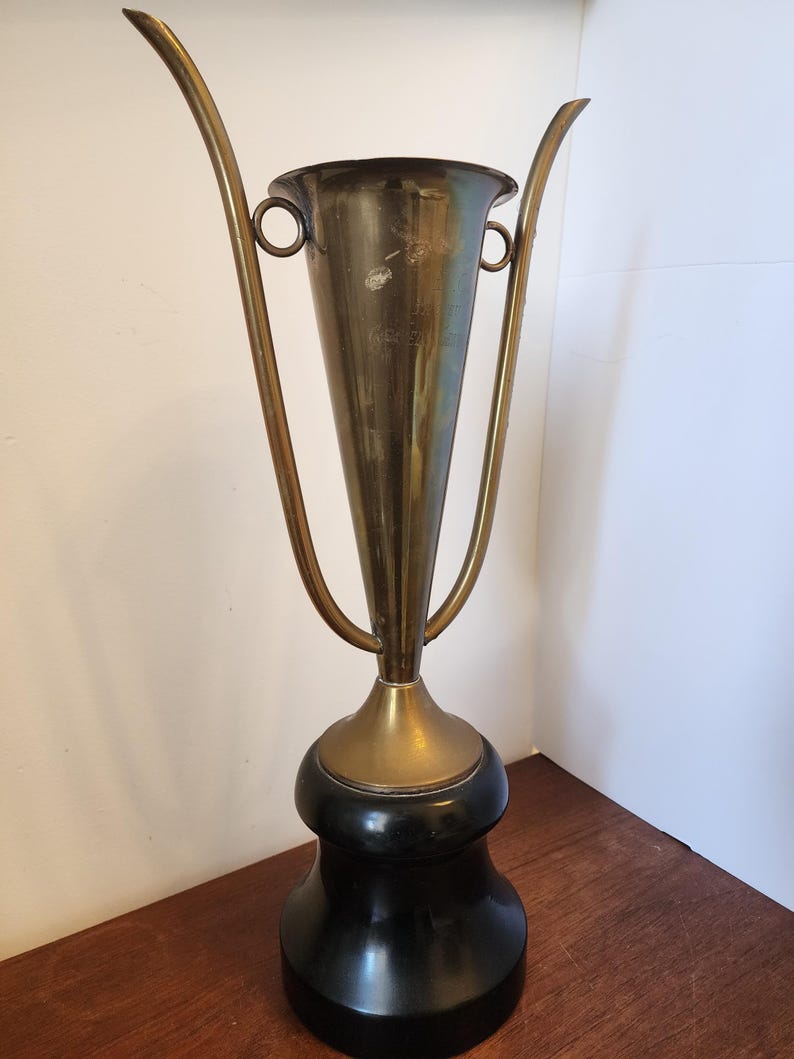 Pode incluir: Uma ta&ccedil;a trof&eacute;u de lat&atilde;o alta com um corpo c&oacute;nico e duas pegas longas curvadas para cima, fixadas a an&eacute;is circulares. O recipiente met&aacute;lico assenta numa base preta lisa e arredondada, criando um contraste entre o metal dourado e o pedestal escuro.