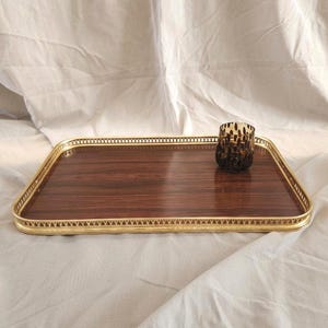 Può includere: Un vassoio da portata rettangolare con una superficie in venature di legno marrone scuro e un bordo color oro metallizzato con un motivo traforato decorativo. Un piccolo portacandele cilindrico in vetro con motivo maculato marrone scuro.