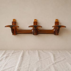 May include: A wall-mounted coat rack made of translucent amber-colored plastic. The rack features three distinct hooks extending from a horizontal bar, with three vertical mounting brackets, each containing a single screw hole for attachment.