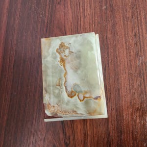 Puede incluir: Una caja rectangular hecha de piedra de &oacute;nix verde pulido, con patrones naturales en espiral en tonos verde p&aacute;lido, blanco y &aacute;mbar intenso. La tapa tiene un acabado liso y brillante con vetas minerales visibles en toda la superficie.