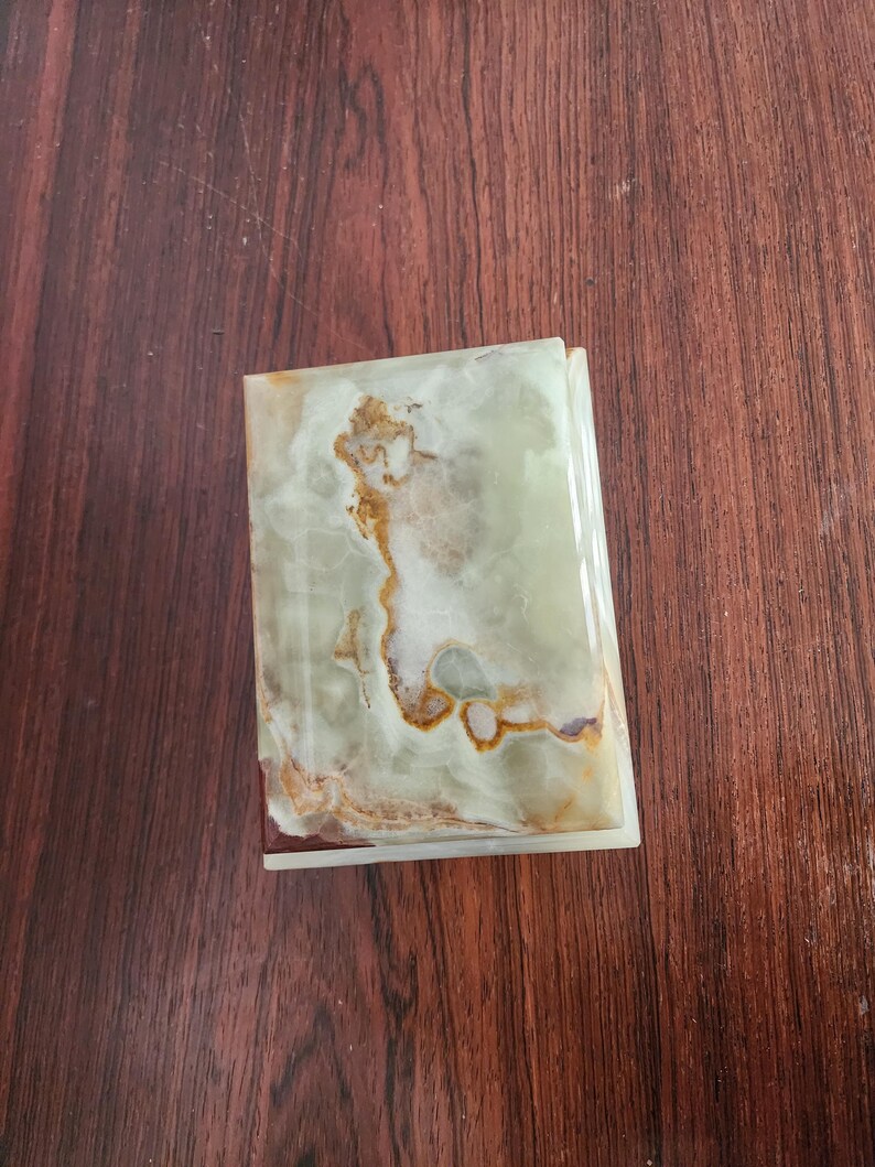 Puede incluir: Una caja rectangular hecha de piedra de &oacute;nix verde pulido, con patrones naturales en espiral en tonos verde p&aacute;lido, blanco y &aacute;mbar intenso. La tapa tiene un acabado liso y brillante con vetas minerales visibles en toda la superficie.