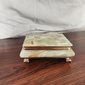 Puede incluir: Una caja rectangular hecha de piedra pulida de color verde y crema con vetas naturales. La caja cuenta con una tapa con bisagras, un borde met&aacute;lico de tono dorado alrededor del centro y cuatro peque&ntilde;os pies decorativos dorados en la base.