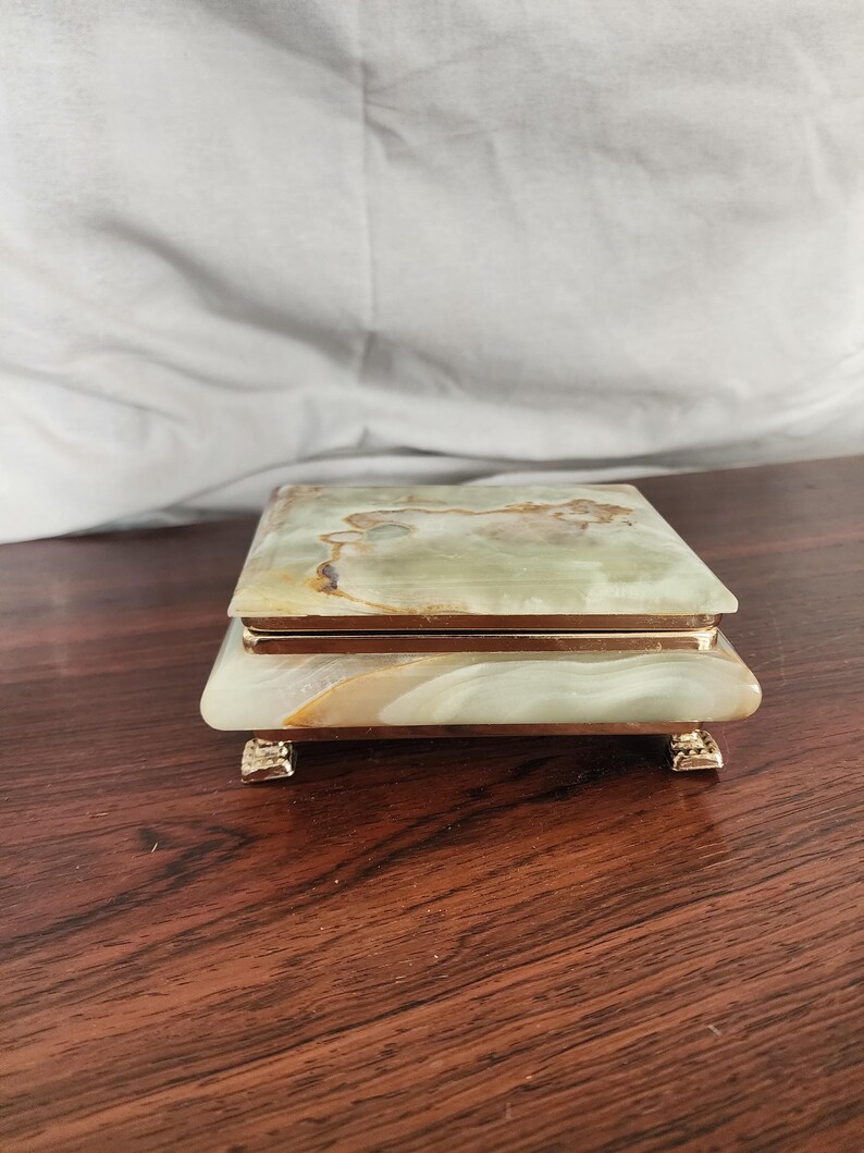 Puede incluir: Una caja rectangular hecha de piedra pulida de color verde y crema con vetas naturales. La caja cuenta con una tapa con bisagras, un borde met&aacute;lico de tono dorado alrededor del centro y cuatro peque&ntilde;os pies decorativos dorados en la base.