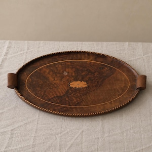 May include: An oval wooden serving tray featuring a dark brown burl wood grain pattern, a rope-textured border, and two side handles. The centre of the tray is decorated with a light-coloured, star-shaped inlay surrounded by a thin circular line.