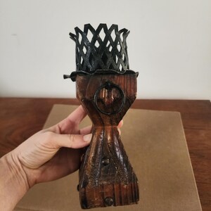 May include: A wooden candle holder featuring a dark brown, textured, carved base with a metal ring accent and a black, lattice-patterned metal cup at the top. The structure is vertical, with a flared base and a narrow middle section.