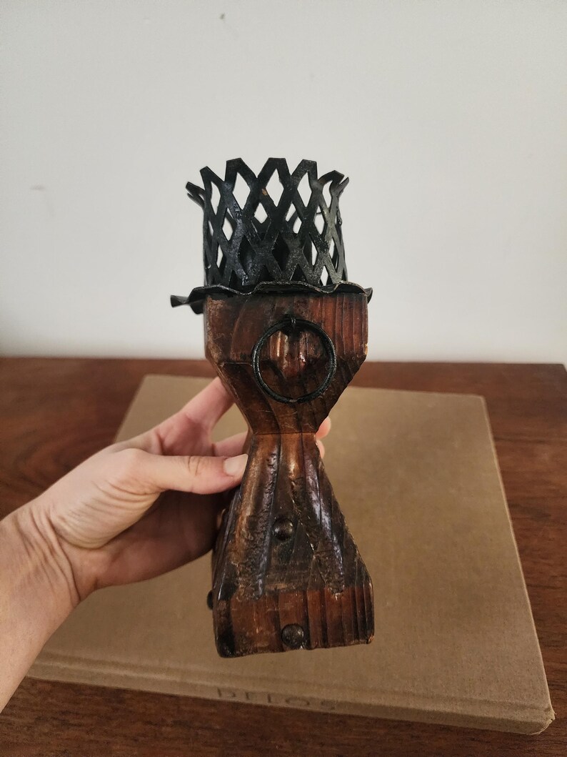 May include: A wooden candle holder featuring a dark brown, textured, carved base with a metal ring accent and a black, lattice-patterned metal cup at the top. The structure is vertical, with a flared base and a narrow middle section.