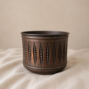May include: A cylindrical copper planter featuring a repeating embossed pattern of vertical fluted panels alternating with rows of small raised beads. The metal surface displays a dark, aged patina with visible oxidation and a smooth rim.