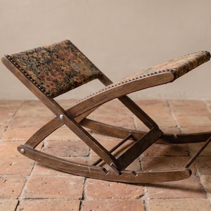 May include: A folding wooden footstool with a dark brown finish and a slanted, upholstered top. The fabric features a multi-coloured floral pattern in muted tones of red, green, and gold, secured with small metal tacks along the wooden frame edges.