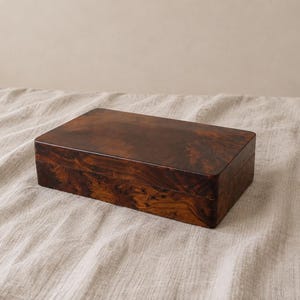 May include: A rectangular wooden box featuring a polished finish with a dark brown and amber swirling grain pattern. The box has a smooth, flat lid and straight sides, showcasing the natural, intricate texture of the wood throughout its surface.