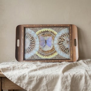 May include: A rectangular wooden serving tray with cutout handles featuring a decorative inlay of three circular patterns. The central design displays a light purple butterfly surrounded by brown and yellow feathers, flanked by two light blue feather circles.