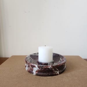 May include: A circular candle holder crafted from dark reddish-brown marble with prominent white veining. The holder features a recessed center designed to securely hold a single, small, cylindrical white pillar candle in an upright position.