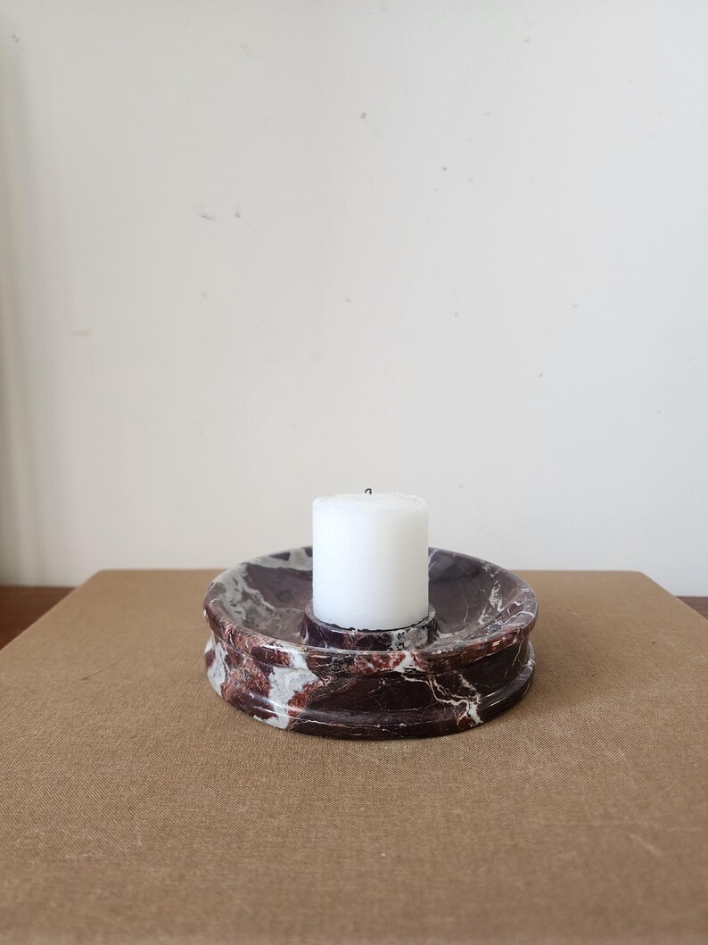 May include: A circular candle holder crafted from dark reddish-brown marble with prominent white veining. The holder features a recessed center designed to securely hold a single, small, cylindrical white pillar candle in an upright position.