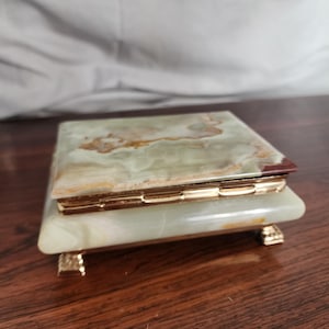 Puede incluir: Una caja rectangular hecha de piedra pulida con bandas verdes y blancas y vetas naturales. La caja cuenta con un marco de metal dorado con bisagras y descansa sobre cuatro peque&ntilde;as patas decorativas de metal dorado en la base.
