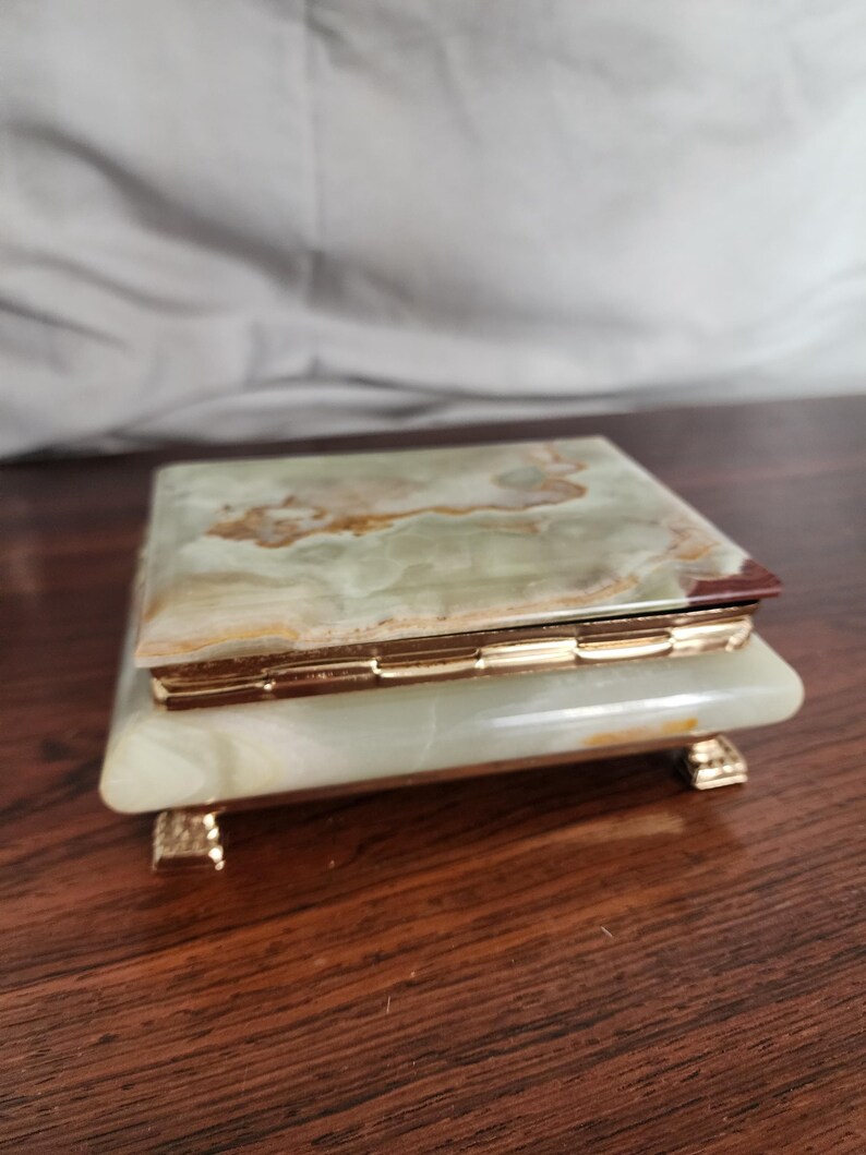 Puede incluir: Una caja rectangular hecha de piedra pulida con bandas verdes y blancas y vetas naturales. La caja cuenta con un marco de metal dorado con bisagras y descansa sobre cuatro peque&ntilde;as patas decorativas de metal dorado en la base.