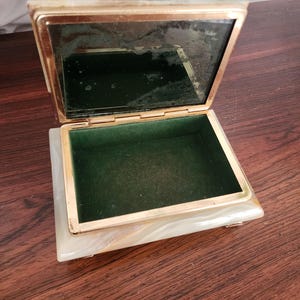 Puede incluir: Un joyero rectangular con exterior de piedra veteada en blanco y crema y marco de metal dorado. La tapa incluye un espejo interior y la base est&aacute; forrada de terciopelo verde oscuro. La caja tiene bisagras y se apoya sobre peque&ntilde;as patas.