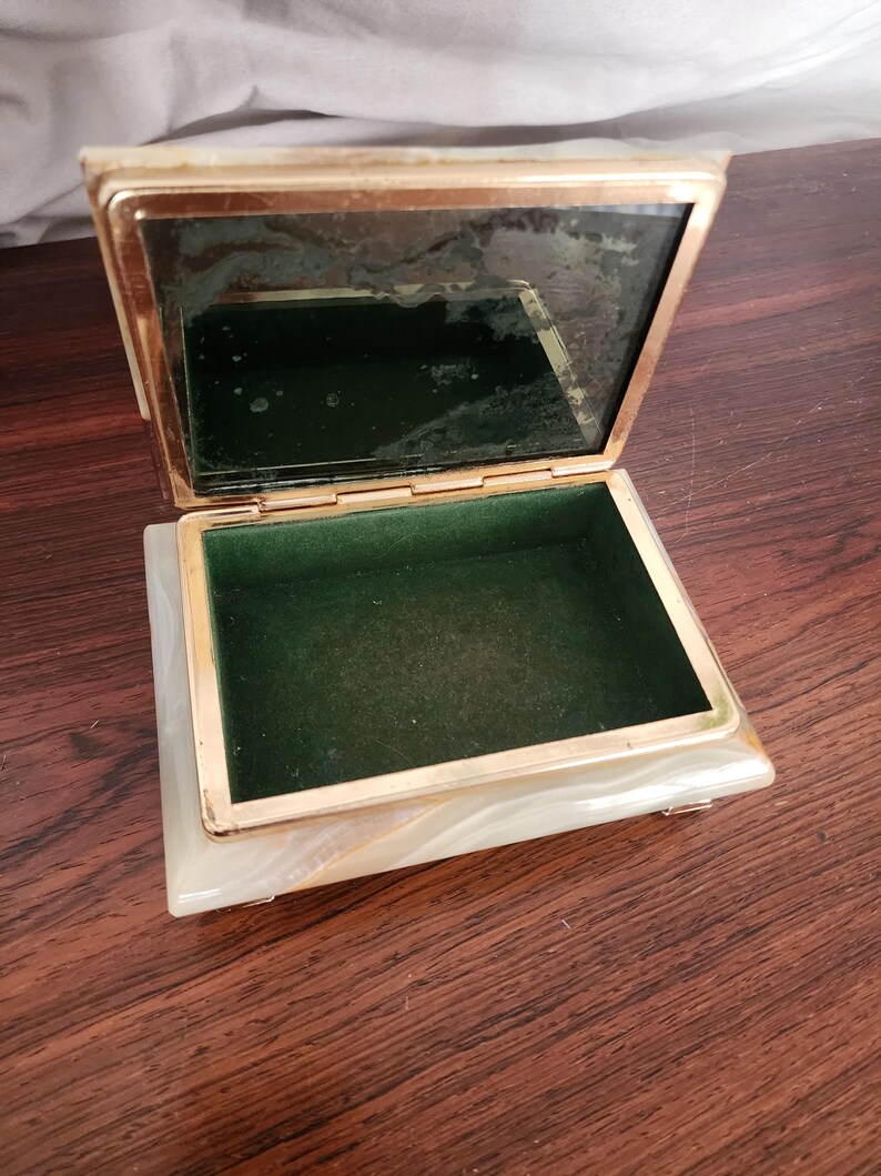 Puede incluir: Un joyero rectangular con exterior de piedra veteada en blanco y crema y marco de metal dorado. La tapa incluye un espejo interior y la base est&aacute; forrada de terciopelo verde oscuro. La caja tiene bisagras y se apoya sobre peque&ntilde;as patas.