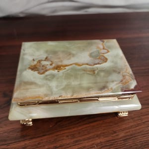 Puede incluir: Una caja rectangular hecha de piedra &oacute;nix pulida en tonos verdes y blancos con vetas marrones naturales. La tapa presenta una superficie lisa, mientras que la base se apoya sobre cuatro peque&ntilde;as patas met&aacute;licas doradas y una bisagra dorada.