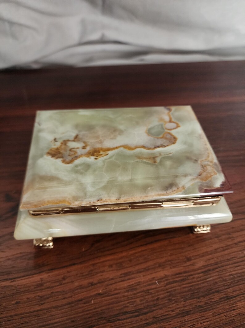 Puede incluir: Una caja rectangular hecha de piedra &oacute;nix pulida en tonos verdes y blancos con vetas marrones naturales. La tapa presenta una superficie lisa, mientras que la base se apoya sobre cuatro peque&ntilde;as patas met&aacute;licas doradas y una bisagra dorada.