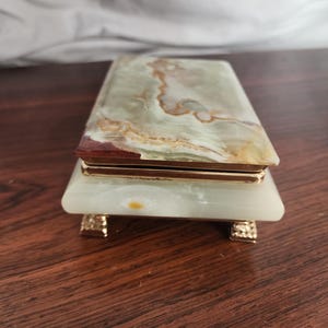 Puede incluir: Una caja rectangular hecha de piedra de &oacute;nice verde pulida con vetas naturales marrones y blancas. La tapa y la base est&aacute;n unidas por un marco de metal dorado, y la caja se apoya sobre cuatro peque&ntilde;as patas de metal dorado texturizado.