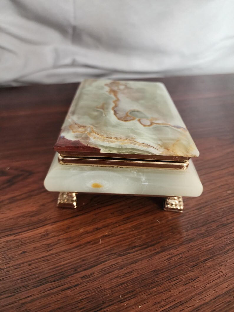 Puede incluir: Una caja rectangular hecha de piedra de &oacute;nice verde pulida con vetas naturales marrones y blancas. La tapa y la base est&aacute;n unidas por un marco de metal dorado, y la caja se apoya sobre cuatro peque&ntilde;as patas de metal dorado texturizado.