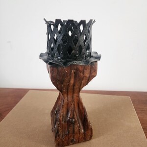 May include: A candle holder featuring a dark brown, carved wooden base with a tapered, textured body and visible metal studs. The top consists of a black metal lattice cage with a scalloped rim, designed to securely hold a candle in place.