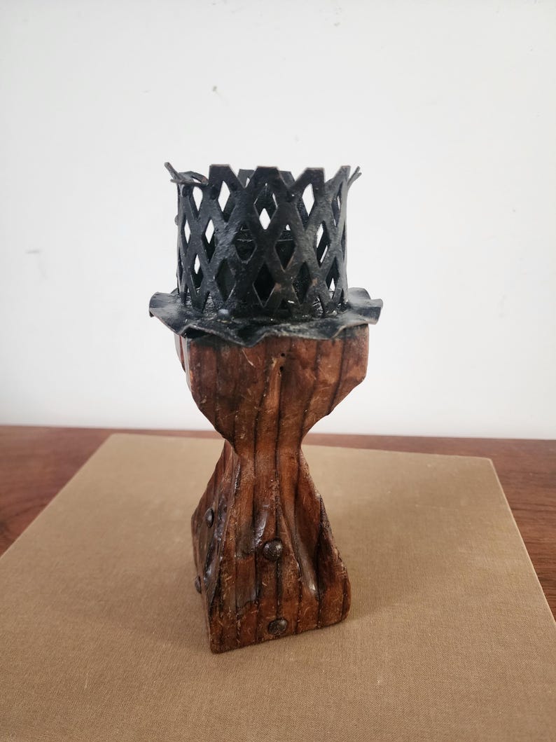May include: A candle holder featuring a dark brown, carved wooden base with a tapered, textured body and visible metal studs. The top consists of a black metal lattice cage with a scalloped rim, designed to securely hold a candle in place.