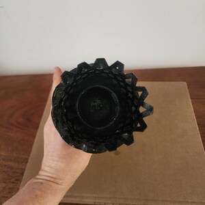 May include: A black metal candle holder featuring a circular base and a decorative, lattice-style rim with a jagged, crown-like edge. The structure is cylindrical, designed to hold a single candle, and displays a dark, matte finish throughout.