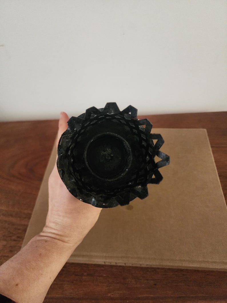 May include: A black metal candle holder featuring a circular base and a decorative, lattice-style rim with a jagged, crown-like edge. The structure is cylindrical, designed to hold a single candle, and displays a dark, matte finish throughout.