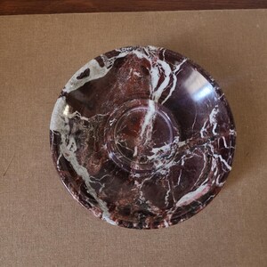 May include: A circular saucer crafted from polished stone featuring a deep burgundy and dark red base color with prominent, irregular white veining throughout the surface. The dish has a smooth, glossy finish and a central recessed circular indentation.