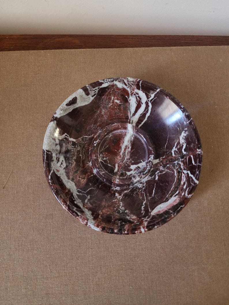 May include: A circular saucer crafted from polished stone featuring a deep burgundy and dark red base color with prominent, irregular white veining throughout the surface. The dish has a smooth, glossy finish and a central recessed circular indentation.