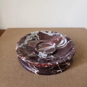 May include: A circular dark red marble tray featuring prominent white veining throughout the stone. The tray has a smooth, polished finish and includes a smaller, raised circular indentation centered within the main basin of the dish.