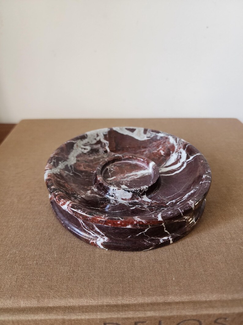 May include: A circular dark red marble tray featuring prominent white veining throughout the stone. The tray has a smooth, polished finish and includes a smaller, raised circular indentation centered within the main basin of the dish.