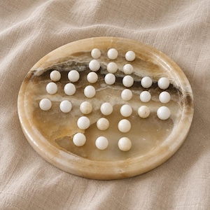 May include: A circular, polished stone game board featuring a natural tan and brown marbled pattern. The board contains a series of small, smooth, white spherical game pieces arranged in a cross-shaped pattern across the indented surface.