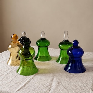 Puede incluir: Una colección de seis pantallas de vidrio para velas en varios colores, incluyendo ámbar, negro, verde claro, verde oscuro y azul. Cada pantalla presenta una base acampanada, una sección media redondeada y un remate de vidrio.