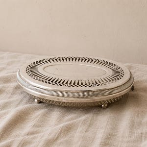 Puede incluir: Una bandeja ovalada de metal plateado con un borde decorativo de celosía calada y un centro sólido. La bandeja tiene un borde estriado y se apoya sobre pequeñas patas redondeadas, mostrando un acabado metálico con detalles recortados.