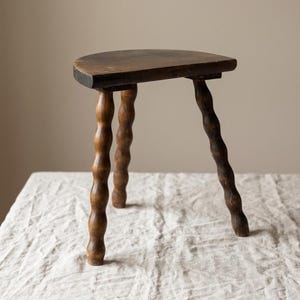 Può includere: Un piccolo sgabello antico in legno con un sedile scuro e semicircolare. Le tre gambe sono intagliate in modo intricato con un design a fuso ritorto. Lo sgabello è posto su una superficie strutturata bianco sporco.