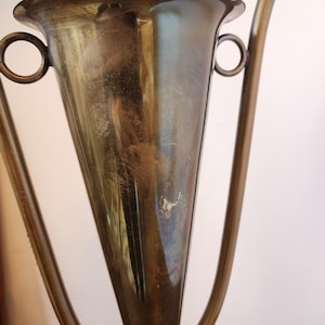 Pode incluir: Uma ta&ccedil;a de trof&eacute;u met&aacute;lica com corpo c&oacute;nico e uma borda larga e alargada. O objeto apresenta duas pegas circulares em forma de anel fixadas nas laterais e uma haste alta e fina. A superf&iacute;cie tem um acabamento polido e reflexivo com sinais vis&iacute;veis de desgaste.