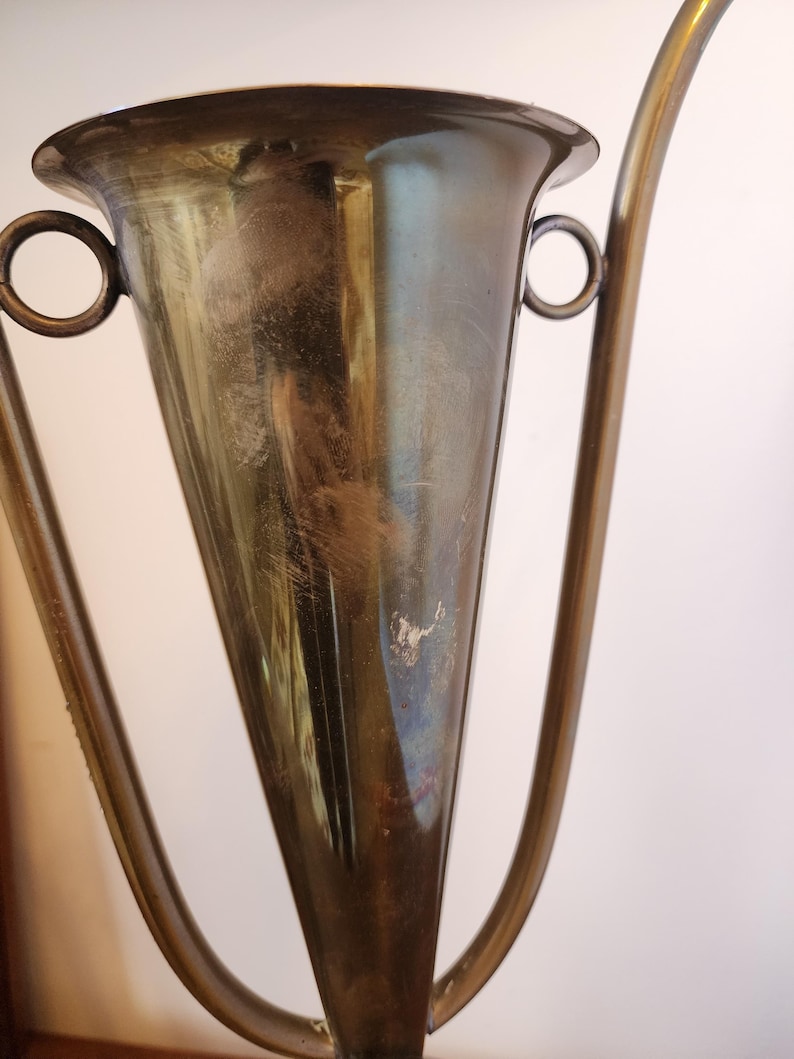 Pode incluir: Uma ta&ccedil;a de trof&eacute;u met&aacute;lica com corpo c&oacute;nico e uma borda larga e alargada. O objeto apresenta duas pegas circulares em forma de anel fixadas nas laterais e uma haste alta e fina. A superf&iacute;cie tem um acabamento polido e reflexivo com sinais vis&iacute;veis de desgaste.